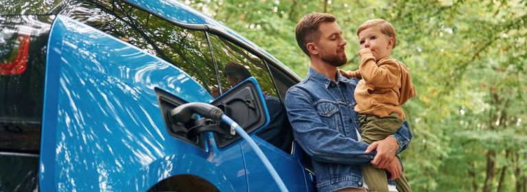 Mann mit Kind am Arm, lehnt am Elektroauto, welches gerade geladen wird - Autoversicherung