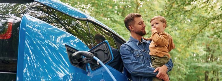 Mann mit Kind am Arm, lehnt am Elektroauto, dass geladen wird - Autoversicherung