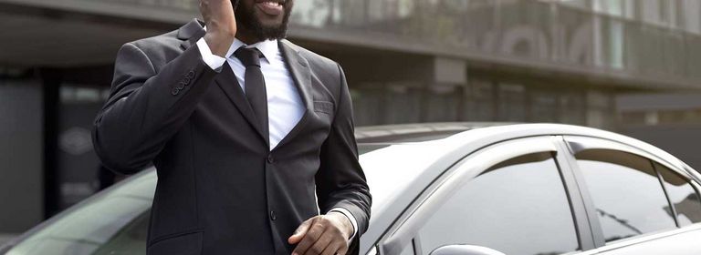 Mann in Anzug steht vor schwarzem Auto und telefoniert
