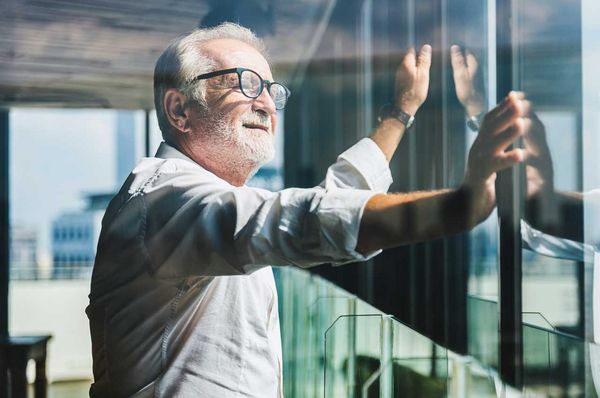 Älterer Mann mit weißen Haaren und Brille steht am Fenster und schaut nach draußen