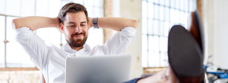 Mann mit Armen hinter dem Kopf, Beine am Tisch und am Schoß den Laptop