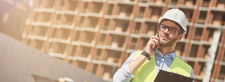 Bauherr steht auf Baustelle und telefoniert, um sich zum Thema Rechtsschutz zu informieren.