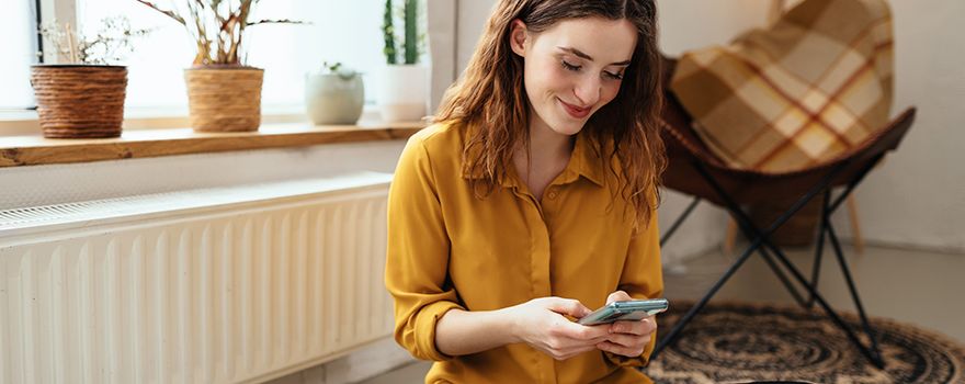 Junge Frau sitzt mit Handy am Boden vor einem Heizkörper