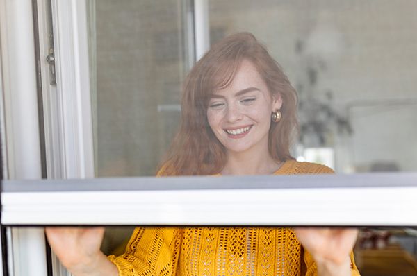 Junge Frau steht am Fenster und schließt das Fliegengitter